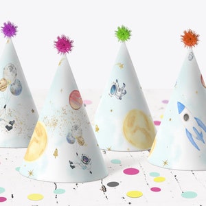 Puede incluir: Cuatro gorros de fiesta azules y blancos con temática espacial. Los gorros presentan astronautas, planetas, estrellas y un cohete. Los gorros tienen pompones coloridos en la parte superior.