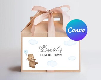 May include: A tan gift box with a light pink ribbon and a white label. The label features a brown bear holding a blue balloon with the number 1. Text on the label reads "Daniel's First Birthday!" and includes blue cloud illustrations.