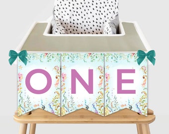 May include: A light blue and white banner with a watercolor design of sea creatures and seaweed. The banner says "ONE" in pink letters and is perfect for a first birthday party.