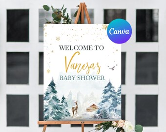 May include: A welcome sign for a baby shower, featuring a winter forest scene with pine trees, a deer, and a cabin. The sign reads "Welcome to Vanesa's Baby Shower" in elegant script and is decorated with gold accents.