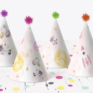 Puede incluir: Cuatro gorros de fiesta rosas y blancos con temática espacial. Los gorros presentan astronautas, planetas, estrellas y un cohete. Los gorros tienen pompones coloridos en la parte superior.