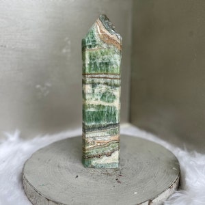 May include: A tall, rectangular green and white stone obelisk with a textured surface. The obelisk has a layered appearance with bands of green, white, and tan. The top is angled. The obelisk is displayed on a round, gray wooden stand.