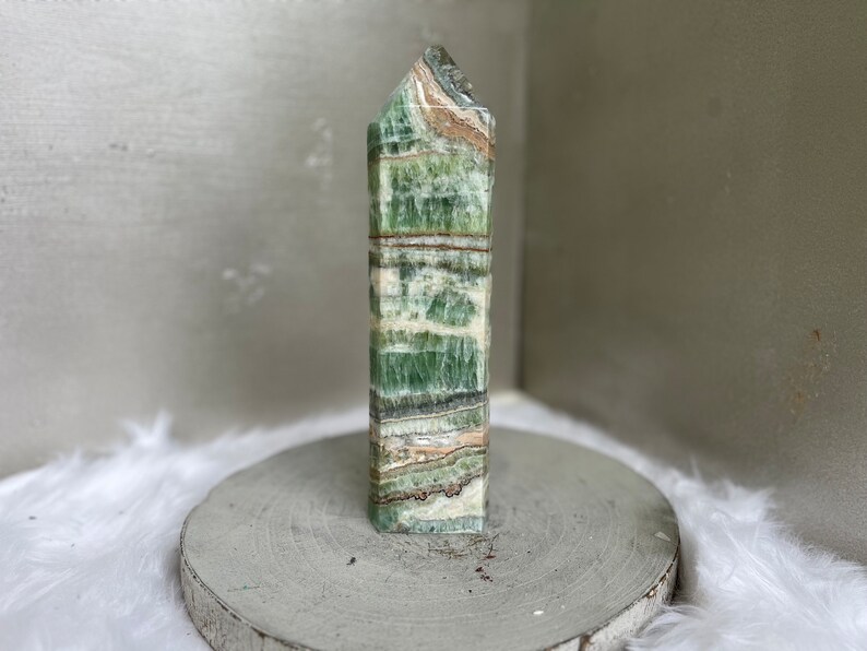 May include: A tall, rectangular green and white stone obelisk with a textured surface. The obelisk has a layered appearance with bands of green, white, and tan. The top is angled. The obelisk is displayed on a round, gray wooden stand.