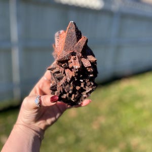 May include: A cluster of reddish-brown crystals, held in a hand, with a rough, textured surface. The crystals have sharp, pointed edges and vary in size, creating a natural, organic form. The background is blurred, with green grass and a white fence.