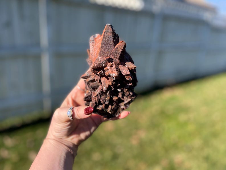 May include: A cluster of reddish-brown crystals, held in a hand, with a rough, textured surface. The crystals have sharp, pointed edges and vary in size, creating a natural, organic form. The background is blurred, with green grass and a white fence.