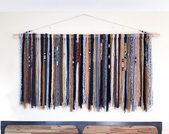 May include: A large, handmade wall hanging featuring vertical strands of yarn in various colors, including black, gray, brown, and pink. The yarn is attached to a wooden dowel and adorned with beads. The wall decor is suspended from a thin string.
