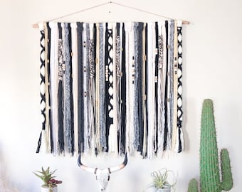 May include: A large, rectangular wall hanging with a wooden dowel. The textile art piece features vertical strands in black, white, gray, and beige, with decorative elements like beads and woven patterns. The piece is designed for home decor.