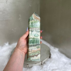 May include: A tall, rectangular green calcite crystal tower with a pointed top. The stone features bands of green, white, and brown. The crystal is held in a hand, showcasing its polished surface and natural patterns. A decorative item.
