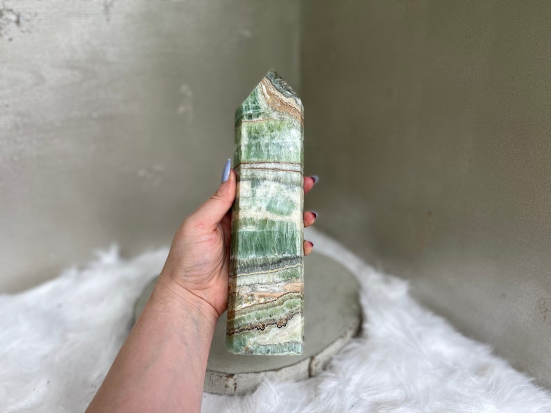 May include: A tall, rectangular green calcite crystal tower with a pointed top. The stone features bands of green, white, and brown. The crystal is held in a hand, showcasing its polished surface and natural patterns. A decorative item.