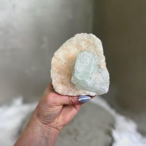May include: A raw, natural crystal formation featuring a large, off-white base with a smaller, light blue-green crystal attached. The crystals have a rough, textured surface. The image is a close-up shot.