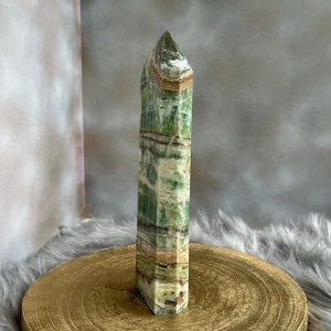 May include: A tall, rectangular green and white crystal obelisk. The crystal has a layered appearance with bands of color. The obelisk is standing on a round, gold-colored wooden surface. The background is a soft, neutral color.