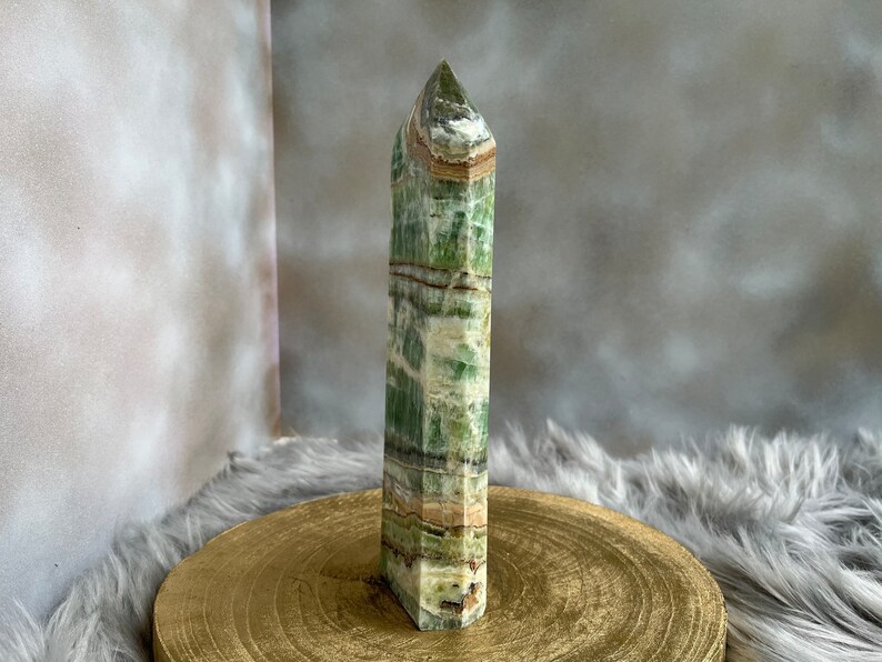 May include: A tall, rectangular green and white crystal obelisk. The crystal has a layered appearance with bands of color. The obelisk is standing on a round, gold-colored wooden surface. The background is a soft, neutral color.