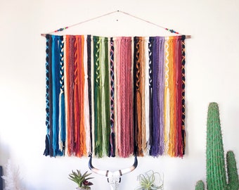 Puede incluir: Un colorido tapiz de pared tejido con hebras verticales en varios colores, incluyendo azul, naranja y morado. La parte superior está unida a una clavija de madera y adornada con cuentas. El tapiz se exhibe contra una pared blanca, con plantas decorativas y una calavera debajo.