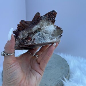 May include: A natural crystal cluster featuring a mix of colors. The crystal has a dark reddish-brown exterior with a lighter, translucent interior. The crystal is held in a hand, showcasing its unique texture and form. The crystal is a natural mineral specimen.