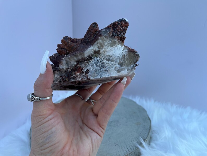 May include: A natural crystal cluster featuring a mix of colors. The crystal has a dark reddish-brown exterior with a lighter, translucent interior. The crystal is held in a hand, showcasing its unique texture and form. The crystal is a natural mineral specimen.