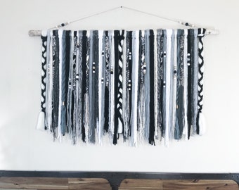 May include: A large wall hanging with a natural wood branch and a variety of hanging yarn strands in black, white, and gray. The strands are adorned with beads and tassels, creating a textured, bohemian-style decor piece.