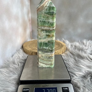 May include: A green and white striped crystal obelisk stands on a digital scale, displaying a weight of 2.700 oz. The crystal has a rectangular base and a pointed top. The scale is on a fluffy, gray surface.