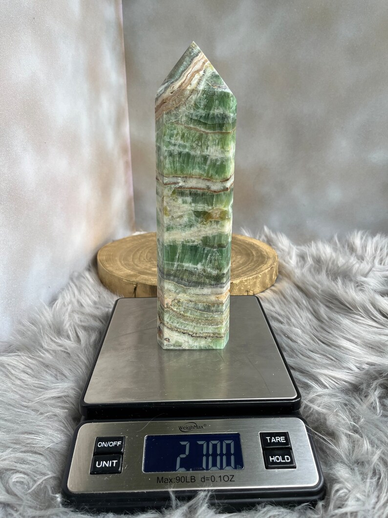 May include: A green and white striped crystal obelisk stands on a digital scale, displaying a weight of 2.700 oz. The crystal has a rectangular base and a pointed top. The scale is on a fluffy, gray surface.