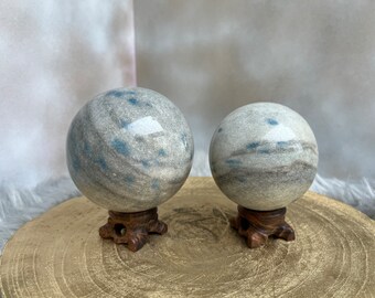May include: Two decorative stone spheres with gray, blue, and brown marbling sit on ornate wooden stands. The spheres are different sizes, with the larger one on the left. The stands are dark brown and have a carved appearance. The spheres are displayed on a gold-colored surface.