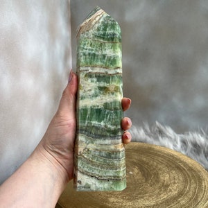 May include: A tall, rectangular green calcite crystal tower with horizontal banding in shades of green, white, and brown. The polished stone has a slightly rough, textured surface and a pointed top. The crystal is held in a hand, with a gold-colored wooden base in the background.