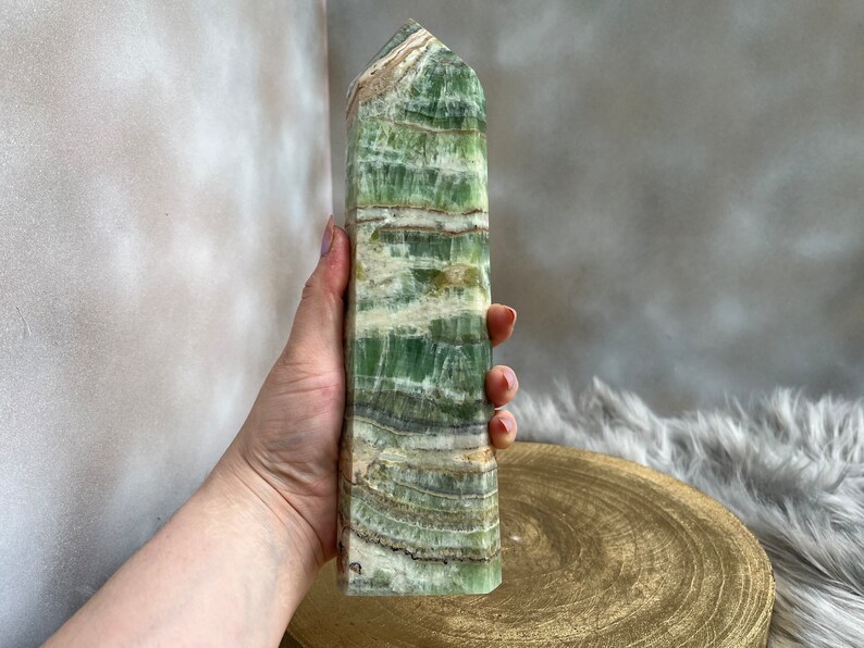 May include: A tall, rectangular green calcite crystal tower with horizontal banding in shades of green, white, and brown. The polished stone has a slightly rough, textured surface and a pointed top. The crystal is held in a hand, with a gold-colored wooden base in the background.