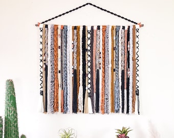 Puede incluir: Un tapiz de pared tejido con una variedad de texturas y colores. La pieza presenta hebras de hilo blanco, negro, marrón y gris, algunas trenzadas, algunas con cuentas y otras con borlas. El tapiz está suspendido de una barra de madera, de unos 60 cm de ancho.