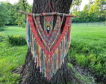 May include: A colorful macrame wall hanging with a wooden dowel. The piece features a V-shaped design with red, orange, blue, and yellow threads, accented with beads. The fringe hangs down, creating a textured, bohemian look.
