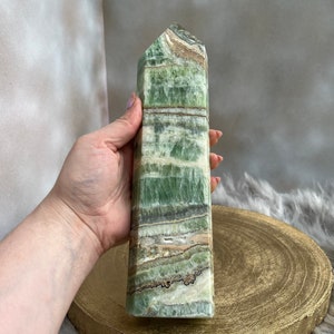 May include: A tall, rectangular green calcite crystal tower with bands of white, brown, and gray. The polished stone has a flat top and is held in a hand. The crystal is displayed on a gold-colored wooden surface.
