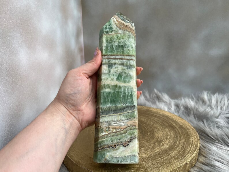 May include: A tall, rectangular green calcite crystal tower with bands of white, brown, and gray. The polished stone has a flat top and is held in a hand. The crystal is displayed on a gold-colored wooden surface.