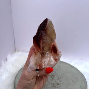 May include: A cluster of natural crystals with a mix of brown, tan, and clear hues. The crystal formation has a rough, textured surface and is held in a hand. The crystals are displayed against a white and gray background.