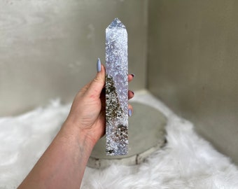 May include: A tall, rectangular crystal point with a polished finish. The crystal features a mix of white, gray, and brown inclusions, creating a unique pattern. The crystal is held in a hand, showcasing its size and shape.