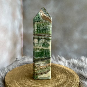 May include: A tall, rectangular green calcite obelisk with brown and white banding. The polished stone is displayed on a gold-colored wooden stand against a neutral background. The obelisk has a pointed top.