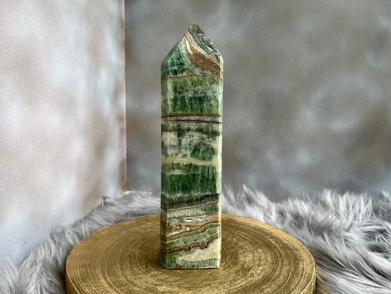 May include: A tall, rectangular green calcite obelisk with brown and white banding. The polished stone is displayed on a gold-colored wooden stand against a neutral background. The obelisk has a pointed top.