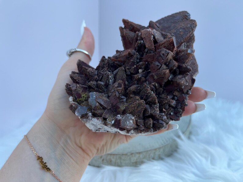 May include: A cluster of dark reddish-brown crystals, possibly a mineral specimen, held in a hand. The crystals have a rough, textured surface and are clustered together. The base of the specimen is a lighter color, possibly white or clear.