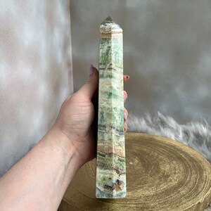 May include: A tall, rectangular green and tan crystal tower with a pointed top. The crystal has a layered appearance with varying shades of green and brown. The crystal is held in a hand.