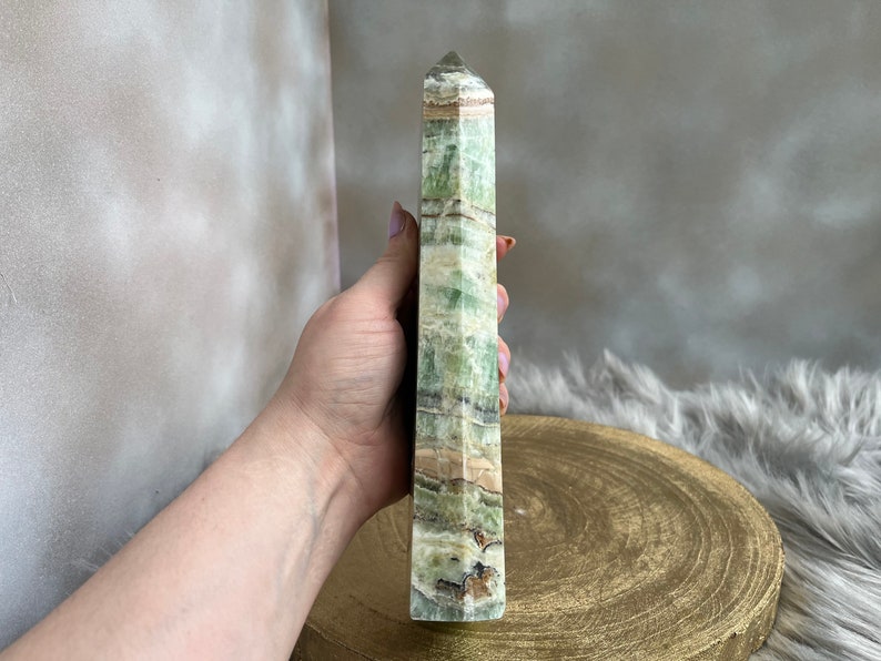 May include: A tall, rectangular green and tan crystal tower with a pointed top. The crystal has a layered appearance with varying shades of green and brown. The crystal is held in a hand.