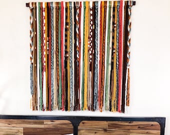 Puede incluir: Un colorido tapiz de pared con hebras verticales de hilo de varios colores, incluyendo marrón, verde, amarillo y rojo. El hilo está sujeto a una barra de madera en la parte superior. Algunas hebras están trenzadas o decoradas con cuentas. Esta pieza de arte textil añade un toque bohemio.