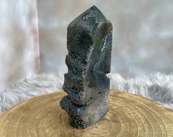 May include: A natural, raw crystal formation with a dark green and gray color palette. The crystal has a rough, textured surface with areas of small, sparkling formations. It is displayed on a gold-colored, round wooden surface.