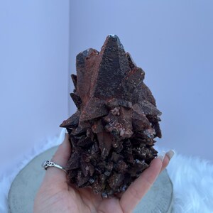 May include: A dark reddish-brown mineral cluster with sharp, crystalline formations. The mineral has a rough, textured surface and is held in a hand. The background is a light purple wall and a white fluffy surface.