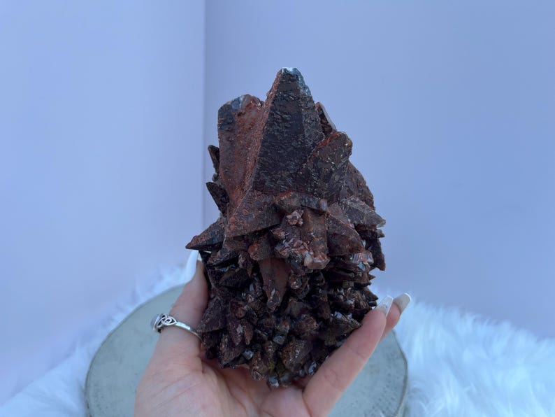 May include: A dark reddish-brown mineral cluster with sharp, crystalline formations. The mineral has a rough, textured surface and is held in a hand. The background is a light purple wall and a white fluffy surface.