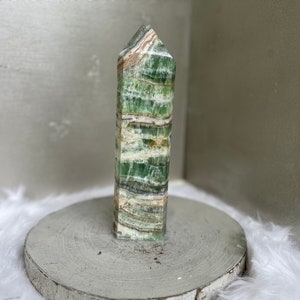 May include: A tall, rectangular crystal tower with a pointed top. The stone features layers of green, white, and brown hues. It is displayed on a round, gray, wooden base, with a white, fluffy surface in the background.