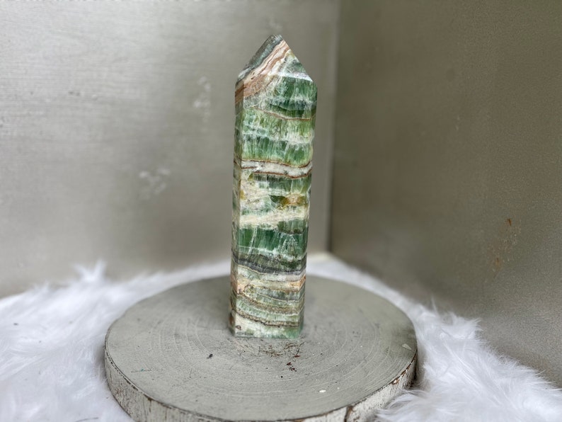 May include: A tall, rectangular crystal tower with a pointed top. The stone features layers of green, white, and brown hues. It is displayed on a round, gray, wooden base, with a white, fluffy surface in the background.