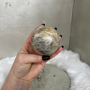 May include: A spherical, polished stone with a swirling pattern of beige, gray, and brown hues. The orb is held in a hand with black nail polish, suggesting a focus on natural elements and decorative objects.