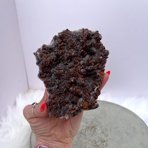 May include: A natural mineral specimen with a rough, textured surface. The crystal formation displays a mix of dark brown and gray hues, with a crystalline structure. The mineral is held in a hand, showcasing its size and detail.