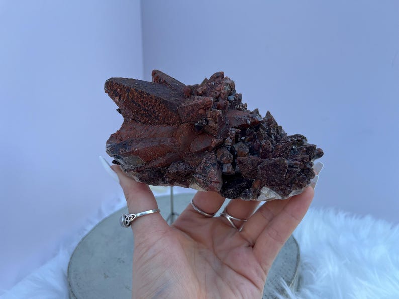 May include: A cluster of dark red quartz crystals with a rough, textured surface. The crystals are a deep reddish-brown color, with some areas appearing almost black. The quartz sits on a white base, and the overall shape is irregular.