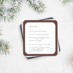 May include: A stack of square Christmas cards with a dark brown border. The cards feature text from Luke 2:10-11, with the words "great joy" in a script font. The cards are surrounded by evergreen branches.