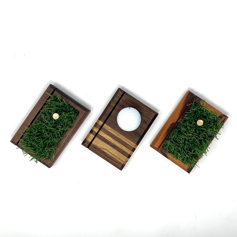 May include: Three wooden golf-themed display trays. Two trays feature a patch of green artificial grass with a golf tee. The third tray holds a white golf ball. The trays are made of dark wood.