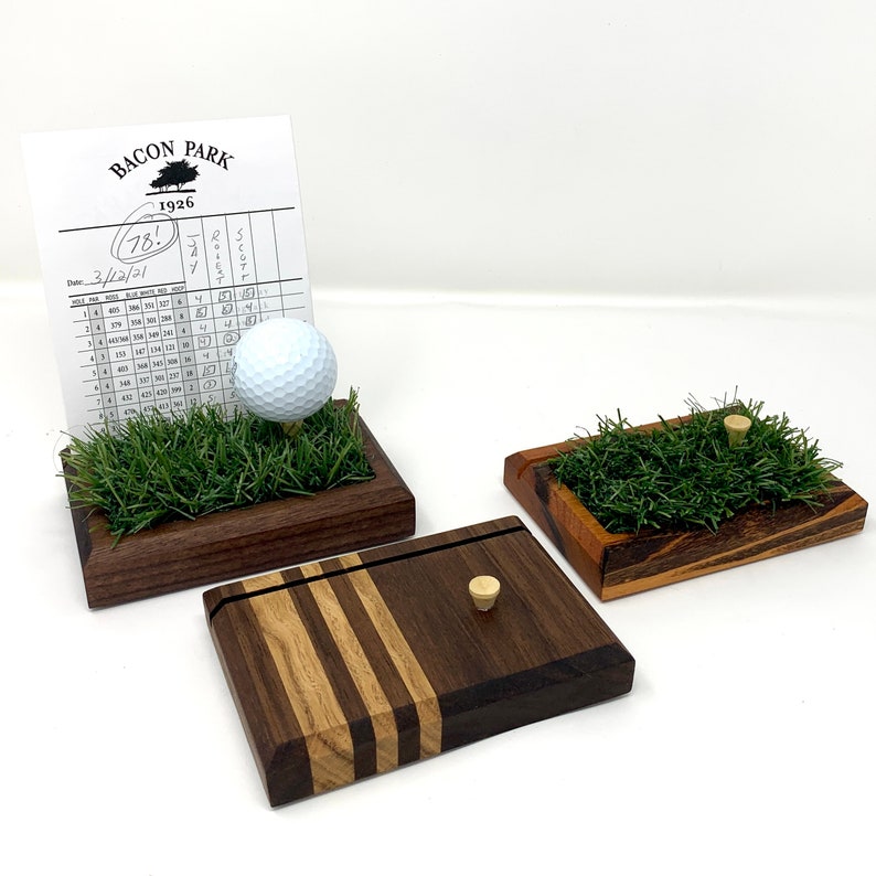 May include: Three wooden golf-themed display pieces. One features a golf ball on a tee in artificial grass, another has a tee in the grass, and the third is a wooden box with a tee. A Bacon Park scorecard is in the background.