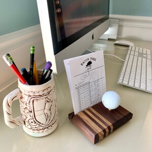 May include: A golf-themed desk arrangement with a scorecard holder, golf ball, and a decorative mug filled with pens and pencils. The scorecard holder is made of wood with a striped design. The mug has a handle and a relief design.