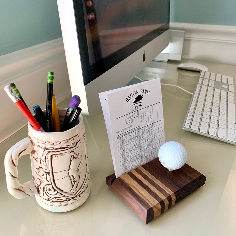 May include: A golf-themed desk arrangement with a scorecard holder, golf ball, and a decorative mug filled with pens and pencils. The scorecard holder is made of wood with a striped design. The mug has a handle and a relief design.